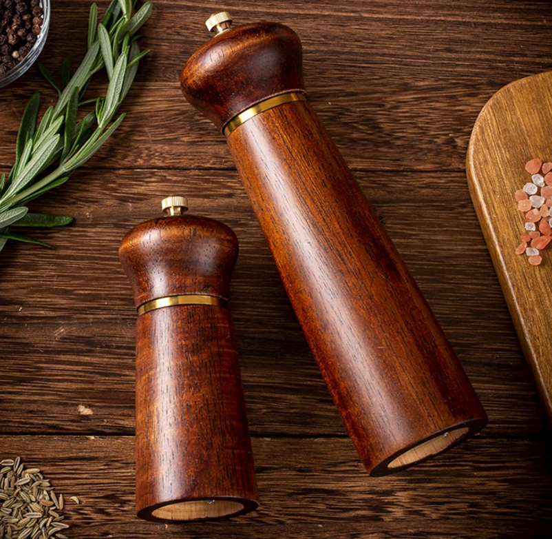 Wooden pepper grinders on a wooden surface with herbs and spices.