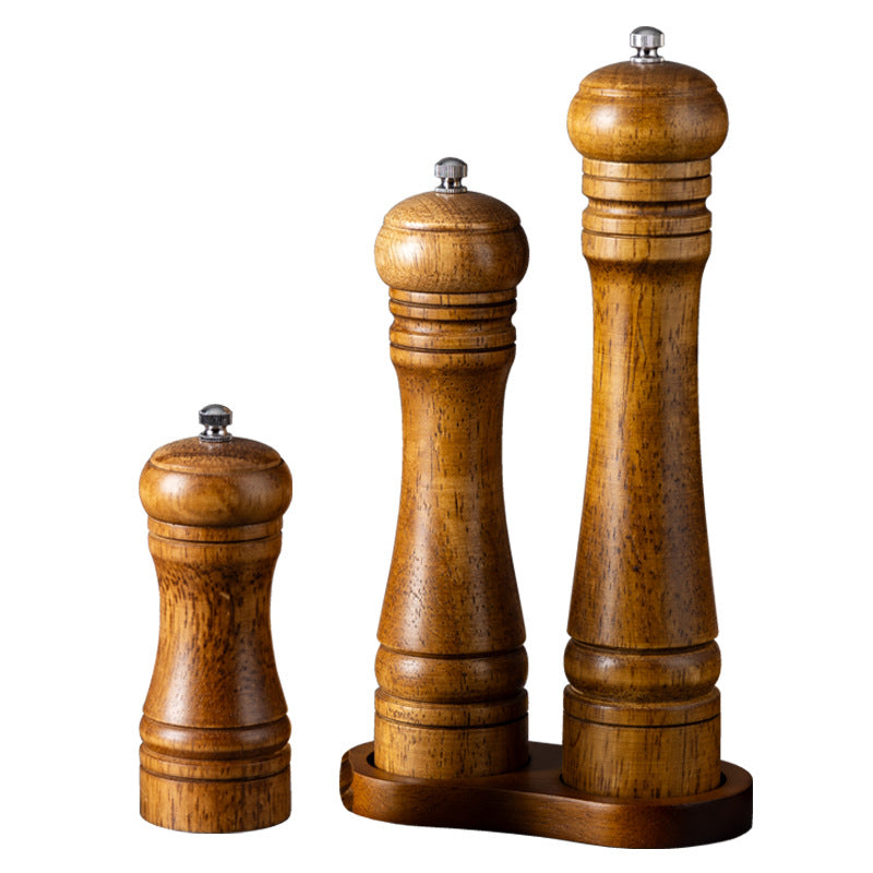 Oak wooden salt and pepper mills of various sizes on a marble background with scattered peppercorns, next to a dish of pasta and a checkered napkin, showcasing the rustic kitchenware