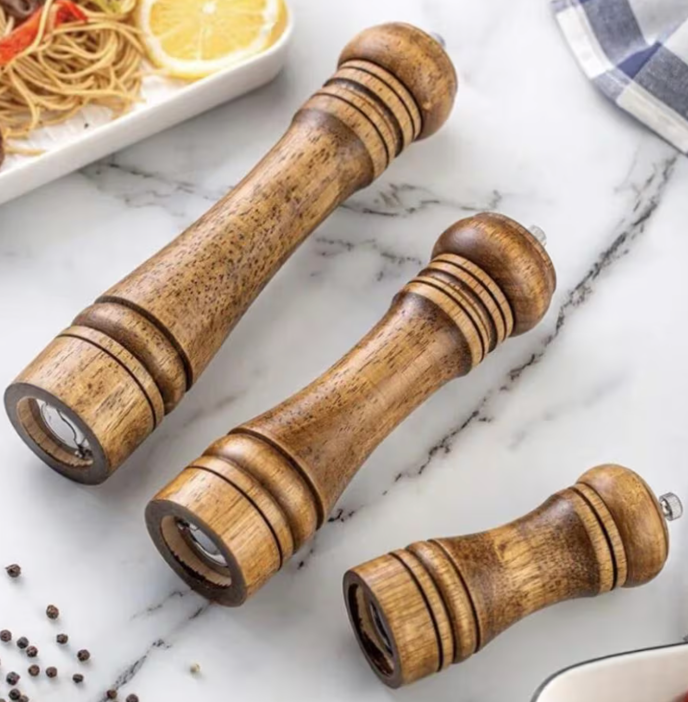 Premium oak wooden salt and pepper mills in various sizes on a marble background with scattered peppercorns. Rustic kitchenware set next to a dish of pasta and a checkered napkin, perfect for home chefs and food enthusiasts. Elegant and durable spice grinders for fresh seasoning, ideal for kitchen decor, dining tables, and gourmet cooking. High-quality wooden peppermill set for a stylish and functional addition to any kitchen or restaurant setting.