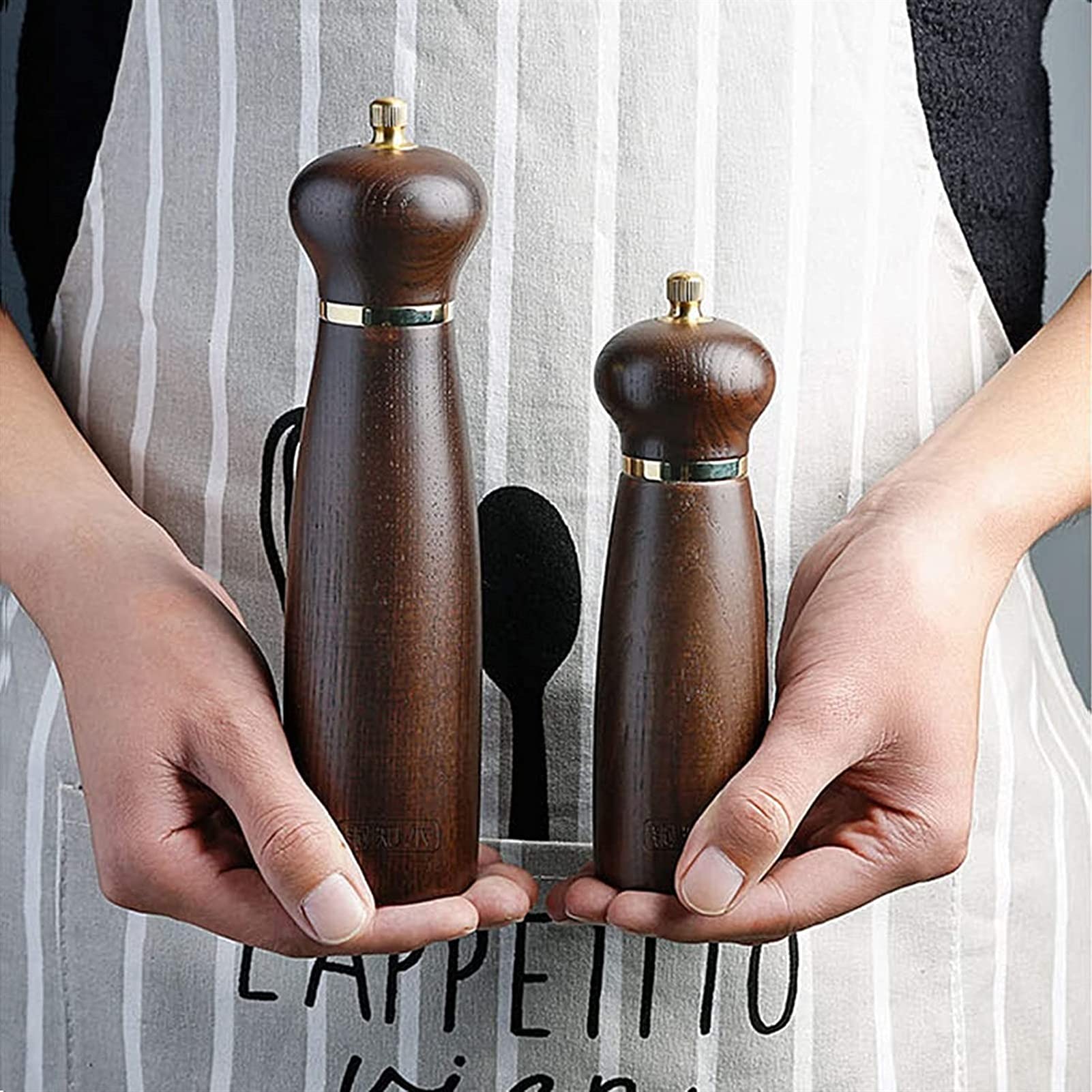 Two wooden pepper mills held by hands with a striped apron in the background.