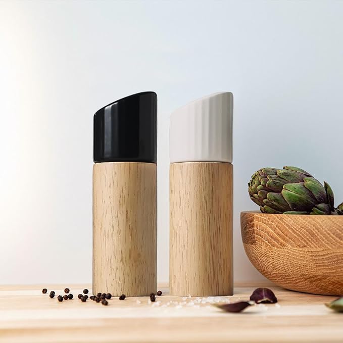 Two modern wooden spice or salt shakers with angled caps stand on a light wood surface against a pale blue background. One shaker has a black cap and the other has a white ribbed cap.