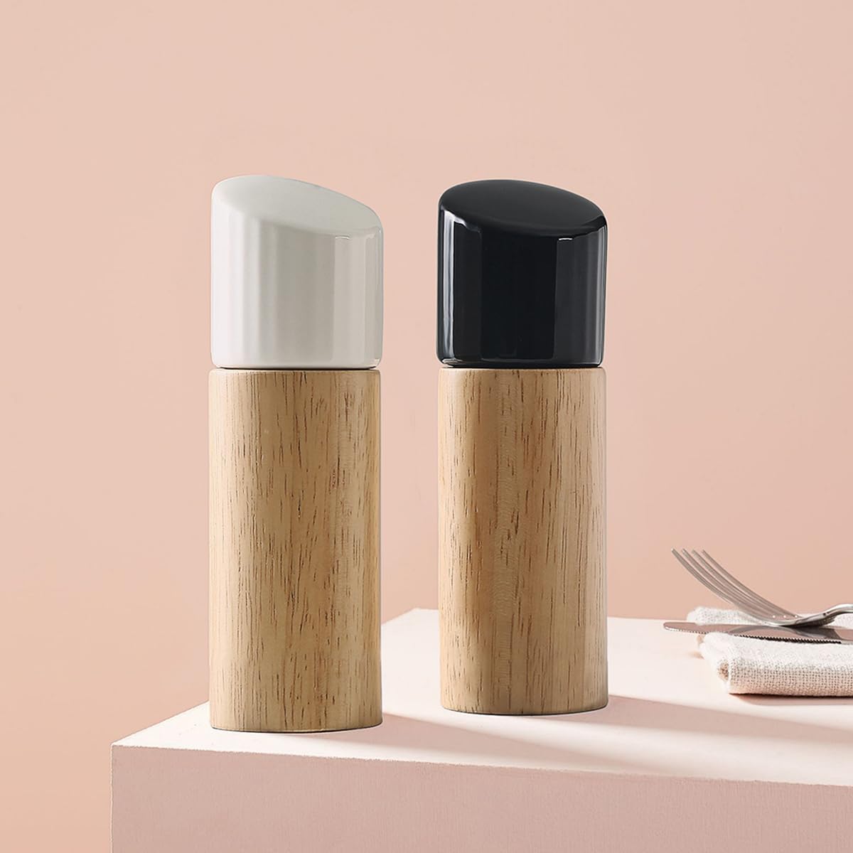 Two modern cylindrical containers with light wood bases, one with a white cap and one with a black cap, displayed on a pink pedestal against a soft pink background.