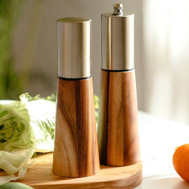 Two wooden and stainless steel salt and pepper mills standing on a round wooden tray, with vegetables and an orange blurred in the foreground and background.