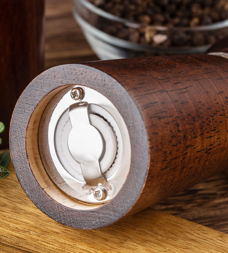 Wooden pepper grinder on a wooden surface with a bowl of peppercorns in the background.