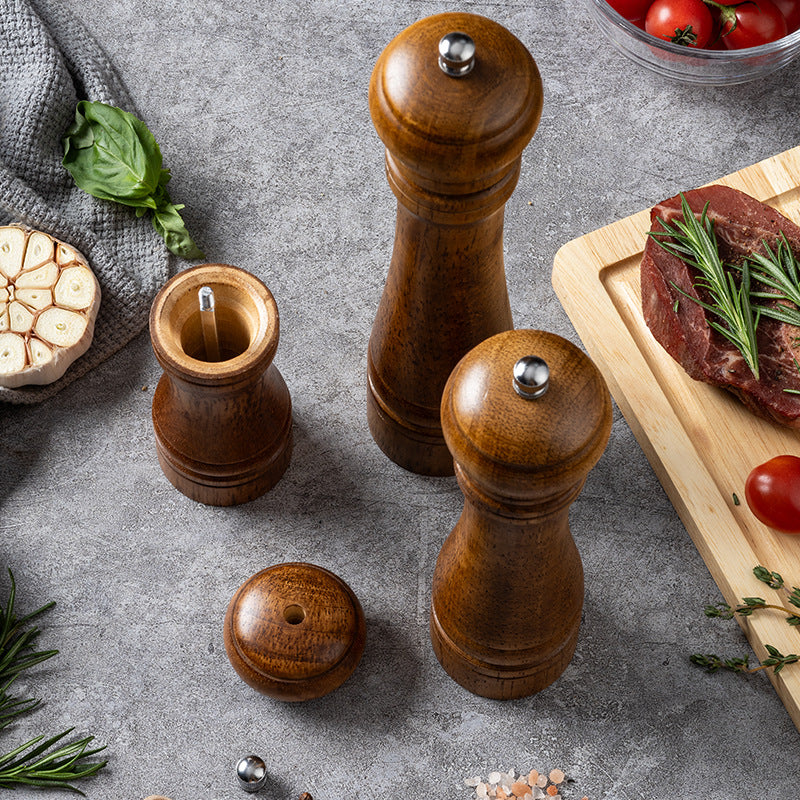 Oak wooden salt and pepper mills of various sizes on a marble background with scattered peppercorns, next to a dish of pasta and a checkered napkin, showcasing the rustic kitchenware