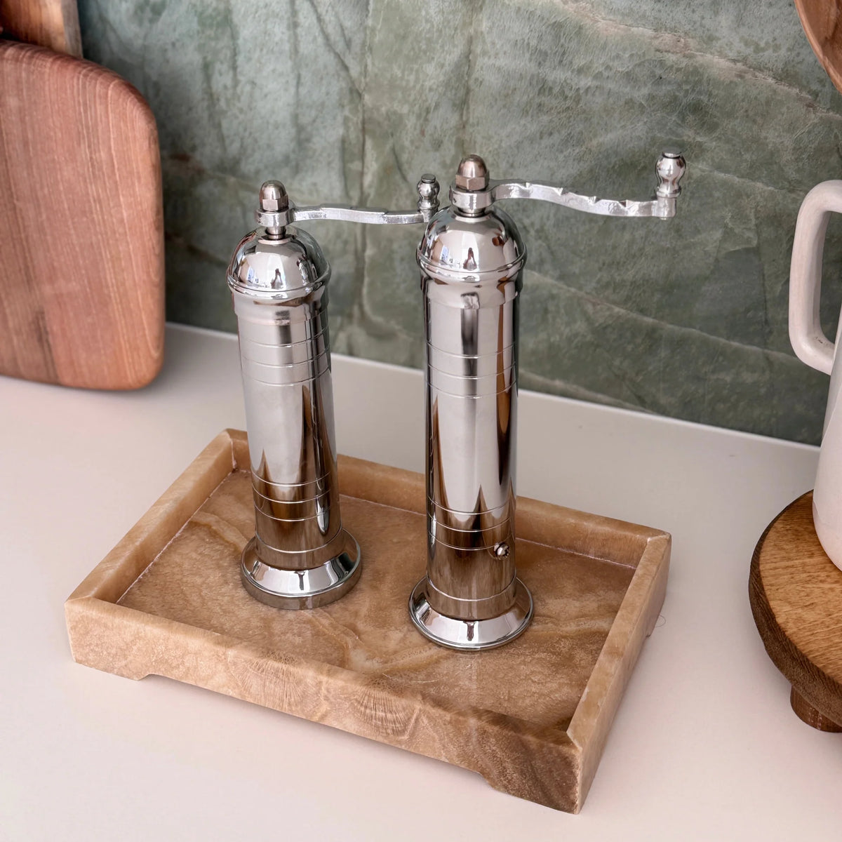 Two silver pepper grinders on a wooden tray with a marble wall in the background.