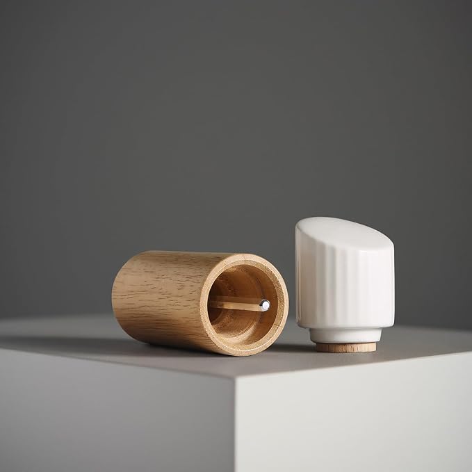 A minimalist wooden and ceramic matchbox set on a light gray pedestal against a dark gray background. One half is a cylindrical natural wood container with a match inside, and the other half is a white ribbed ceramic cap with a small wooden base.
