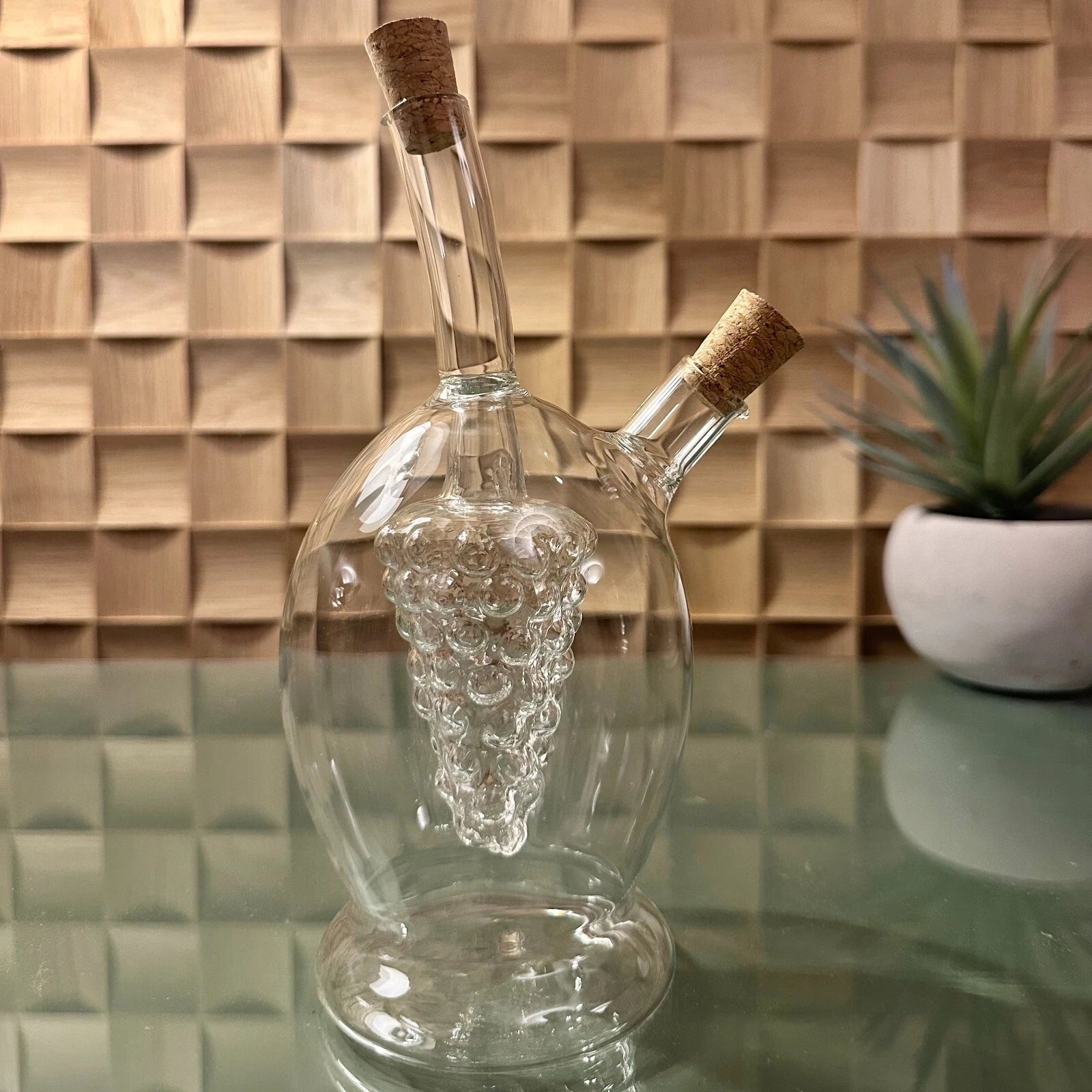 Clear glass decanter with grape design on a reflective surface with a wooden wall background