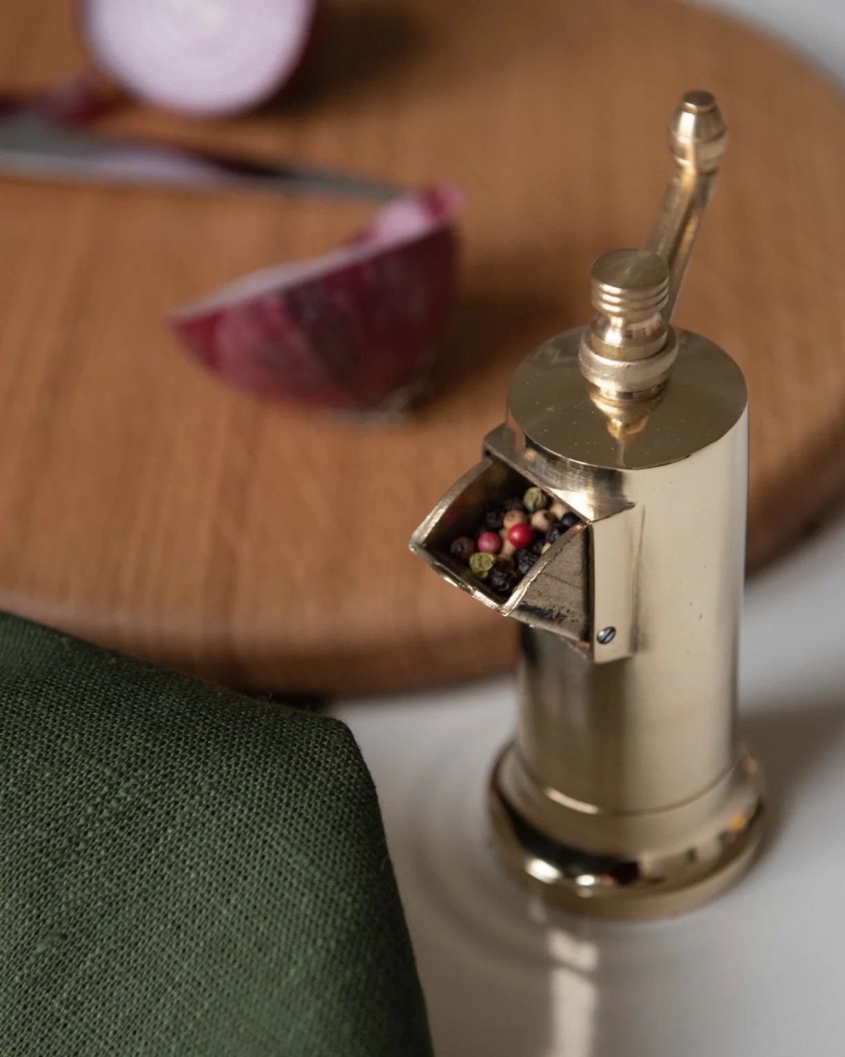 Gold-colored Atlas Pepper Mill with a side crank handle, placed on a marble countertop with ground black pepper sprinkled around.